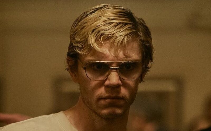 Evan Peters, dressed as Jeffrey Dahmer, wears thick-rimmed glasses and a blank expression in a yellow-tinted still from the series.